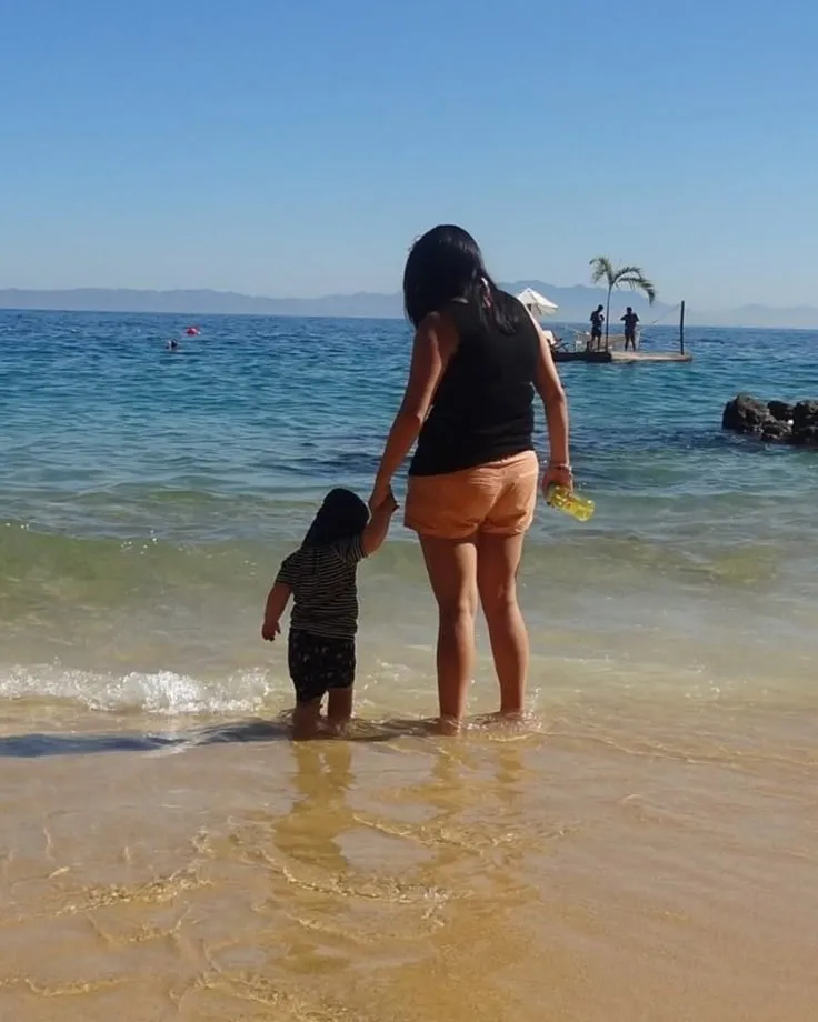 Kate paddling at the water's edge on a sunny beach, holding a little one's hand